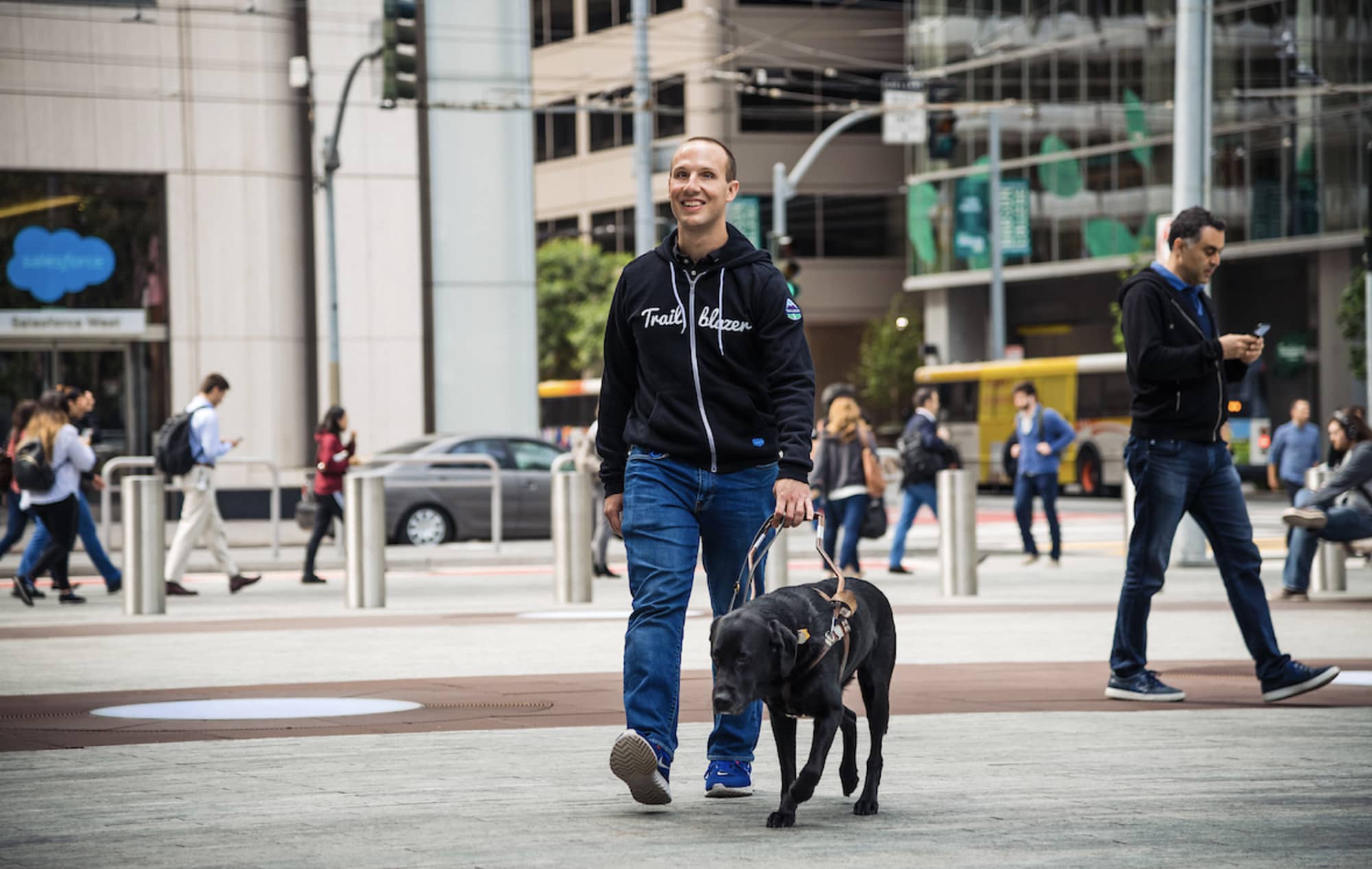 A smiling Salesforce employee wearing a Trail Blazer sweatshirt and walking with his guide dog in the city near Salesforce HQ.