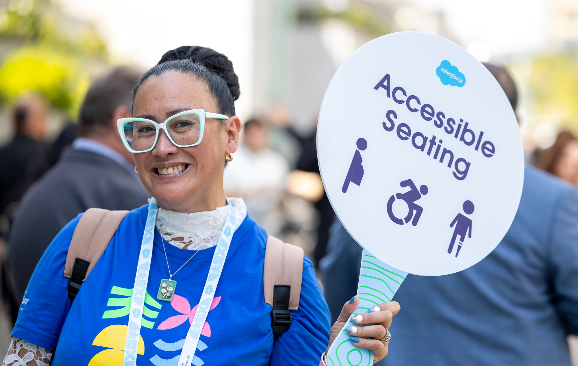Smiling woman in white-framed glasses holds accessible seating sign with accessibility icons at Dreamforce.